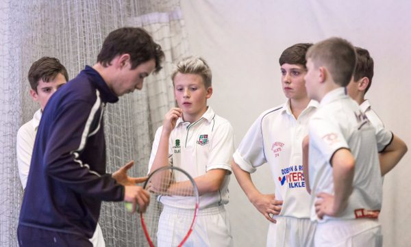 a cricket coach in blue tracksuit talks to a semi-circle of five boys at indoor cricket nets