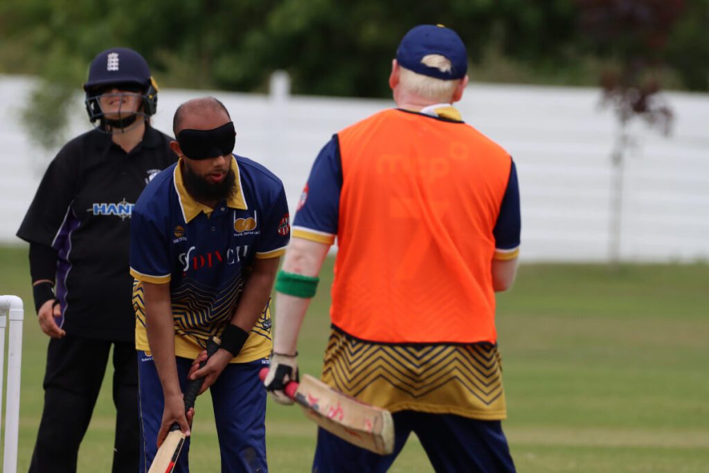 A photo of a Bradford VICC batter who is a total or B1 waiting to play a shot while his runner in an orange bib stands nearby. behind is a B1 Yorkshire Vikings fielder with black wristband and England helmet.