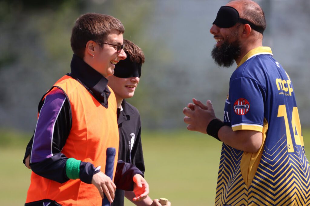 A Yorkshire Vikings batter with black wristband and black eyemask is led by a runner in orange bib and green wristband to the the other end of the pitch, passing a Bradford VICC player.