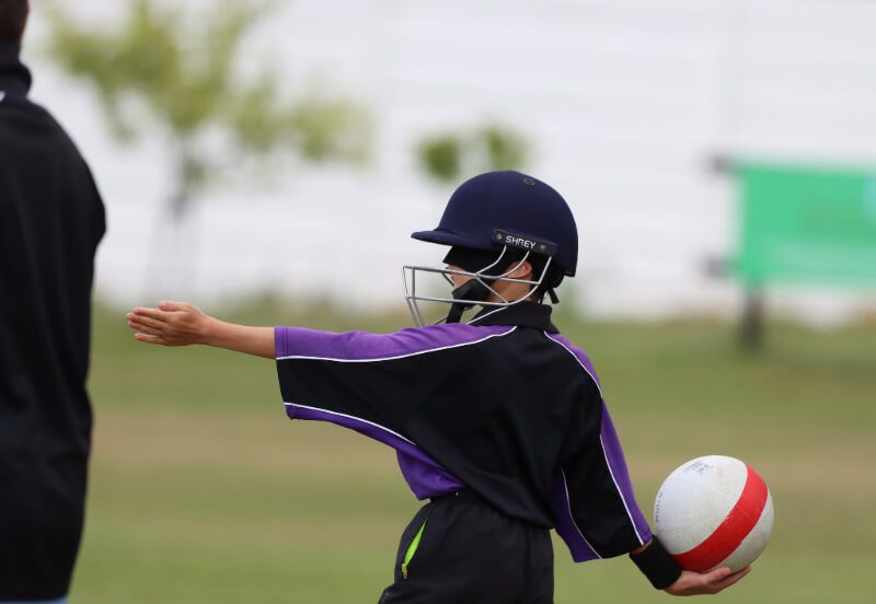 A young Yorkshire Vikings bowler, in black and purple shirt, aims with his left hand straight out in front while holding the ball behind him with the other hand. He wears a blue Shrey helmet and his black eyemask to denote his sight category.