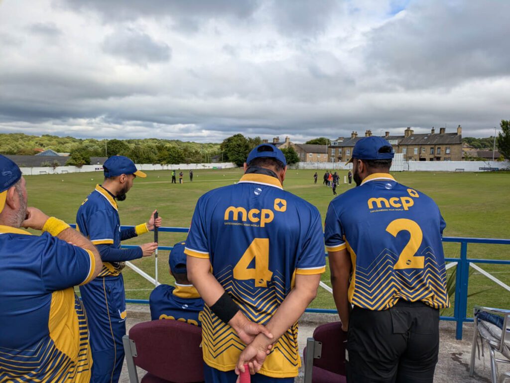 The backs of the Bradford VICC cricketers who are sitting and standing as they watch the action in the middle and wait their turn to bat.